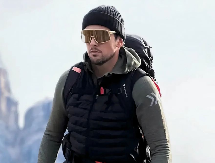 Man in a black beanie and gold NEON OPTIC glasses, wearing a black winter vest and a green-grey thermal top