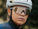 Kobieta w białym kasku rowerowym i złotych okularach sportowych NEON OPTIC, ubrana w granatową koszulkę kolarska